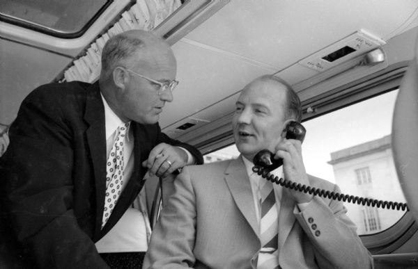 Mayor Ivan Nestingen (seated) using a mobile phone as Arthur Genet, president of the Greyhound Corp. looks on. They are inside a Greyhound Scenicruiser bus, its interior converted into a "traveling office" for Mr. Genet's use. Arthur Genet (of Chicago) was at the beginning of an eight-day inspection tour of Greyhound territory in four states and three Canadian provinces.