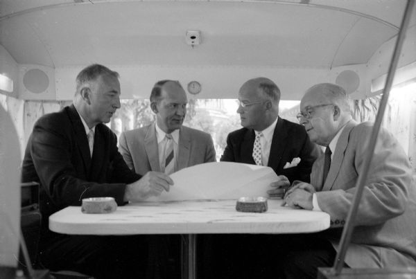 Four men seated at a table inside a customized Greyhound Scenicruiser bus. They are: Arthur Genet (center right), president of the Grayhound Corporation; Mayor Ivan Nestingen (center left); Leroy Luberg (far left), executive secretary to Gov. Vernon Thomson; Atty. Glenn Stephens (far right), council for Northland Greyhound. Ashtrays are on the table where they are looking over papers. 