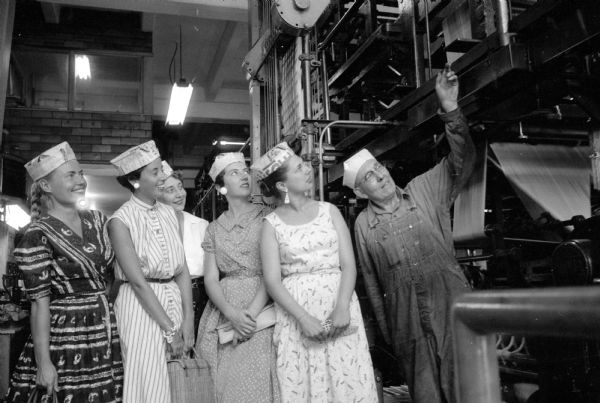 Five women wearing folded newspaper hats while viewing a large printing press on a tour at Madison Newspapers Inc. The five women from the advertising department at Manchester's Department Store are (left to right): Ron Duckwitz, Sally Mohar, Dorothy Schneider, Dorene Goeman, and Yvonne Mamuls, advertising director at Manchester's. At right, is pressroom foreman Mike Luloff, who is showing the women around.