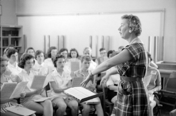 Darleen McCormick, music teacher from Central High School, is shown directing a chorus at a three-week music clinic held at the University of Wisconsin. Students are sitting in rows, are holding sheet music.