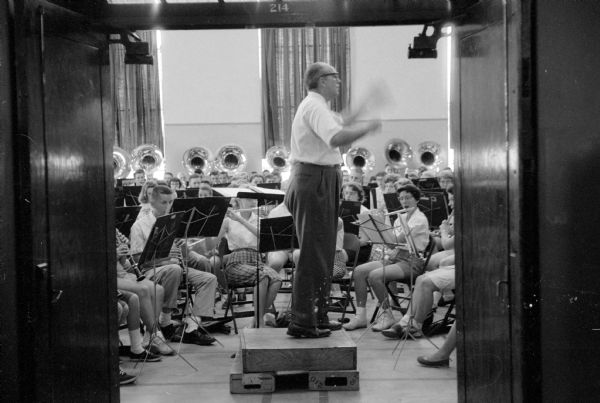 Conductor Milburn Carey from Enid, Oklahoma, is conducting the youth orchestra at the University of Wisconsin Summer Music Clinic. Seen through a doorway, the is standing on a small platform surrounded by students playing flutes, clarinets, and tubas.