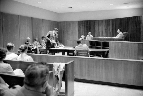Inquest held in the death of Police Sergeant Ralph Nedderman caused by a traffic light violation by driver Alphonse Reiter (23). Shown in the court room are Coroner Michael Malloy (upper right), presiding at the inquest; Alphonse Reiter, on the witness stand answering questions; District Attorney Joseph Bloodgood (standing); and jurors, seated along the left wall. The jury found Reiter guilty of ordinary negligence.      