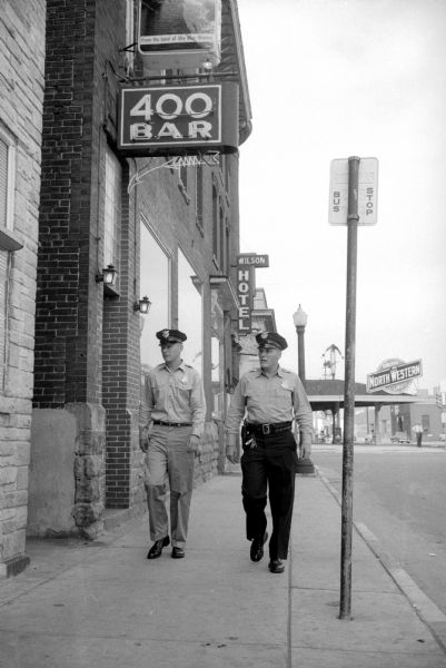 Madison recruit policeman Franklin Oswald (left) learns how to operate on the beat by walking with veteran patrolman Eugene Buchanan (right) on Post 33, the "Midway" or "Skid-Row" beat on East Wilson Street. Above them is a sign that reads "400 Bar" and in the background is the Wilson Hotel and a North Western gas station. 