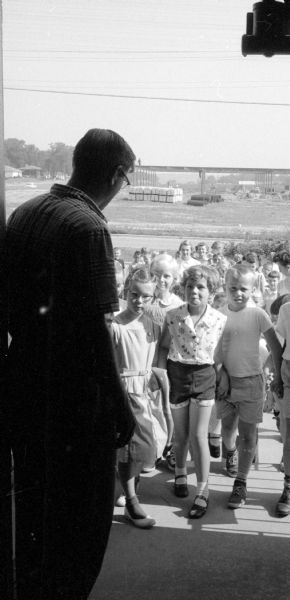 One hundred children attend summer Bible school at the Midvale Community Lutheran Church on 4329 Tokay Boulevard. Shown is the church pastor Reverend Sig G. Sandrock (left) greeting the children. The first children entering the church are (left to right): Jill Mohaupt (4413 Rolba Lane); John Meissner (42 Merlham Drive); and Paul Relerson (510 Clifden Drive). There is an open field across the street in the distance. 