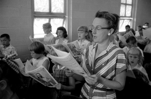 Children attend the summer Bible school session at Midvale Community Lutheran Church at 4329 Tokay Boulevard. Shown singing are 1st-grade teacher Mrs. P. J. Curran (541 Togstad Glen) and students Marcia Sandrock (of Fairbanks, Alaska) in the center and Linda Bogle (4518 Wakefield Street) to her left. All are holding song books with the image of Jesus Christ on the cover.
