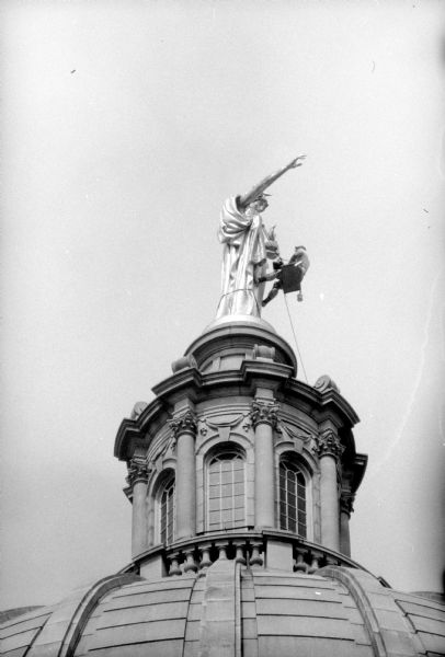 "Wisconsin" - the statue atop the State Capitol building's dome. A painter is hanging from the statue's neck by a climbing rope, pulleys, and a bench while applying gold paint.    