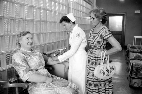 Mrs. J. Leonard Brook, from Shepley, England, is shown having her blood pressure checked by Joan  Dufresne, a Red Cross nurse. Mrs. Brook's hostess (and distant relative), Mrs. Ray S. Owen (right) watches the procedure.