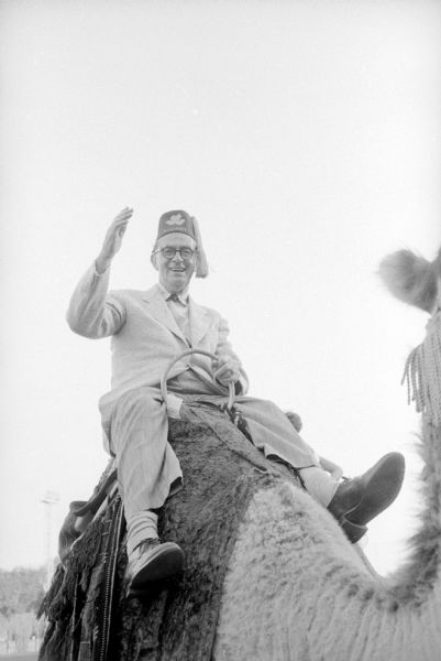 Journalist Joseph "Roundy" Coughlin is riding a camel at the Shrine-Knights of Columbus benefit show at Breese Stevens Field. He was honored for his work with handicapped children. He is wearing a Shriner's fez and waving to the photographer. 