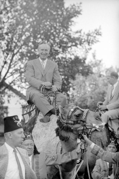 Wisconsin Governor Vernon W. Thomson atop a camel at the Shrine-Knights of Columbus benefit show at Breese Stevens Field. Shriners in their fezzes are below guiding him and other riders. The camel has a decorative harness and is frothing at the mouth.