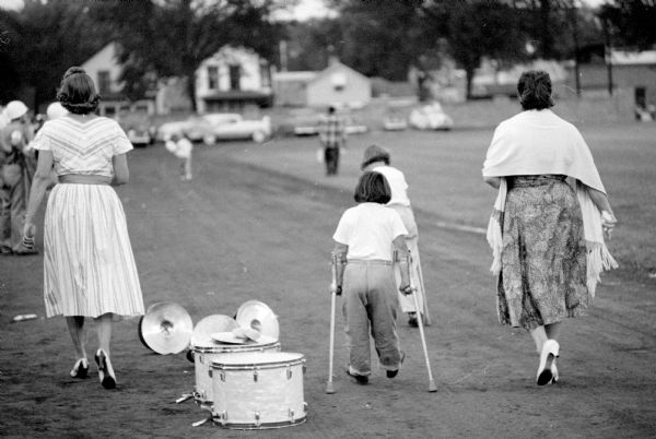 A woman and a disabled child with crutches leaving the field after the Shrine-Knights of Columbus benefit show at Breese Stevens. They are walking past a drum set from the marching band.