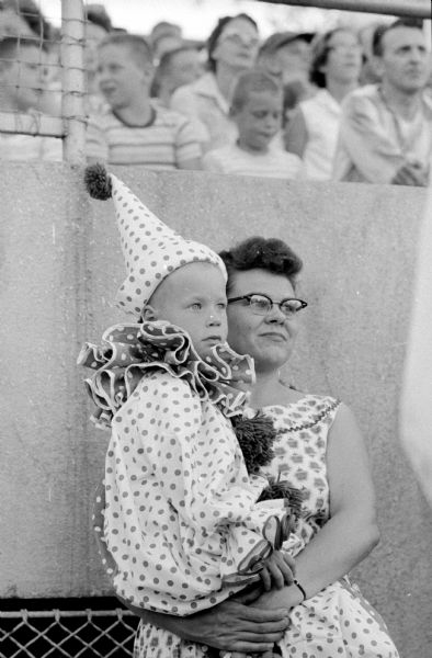 Mark Struve, in a clown suit, is being held by his mother, Mrs. Edward Struve, at the Shrine-Knights of Columbus benefit show at Breese Stevens Field. They are standing below the bleachers.