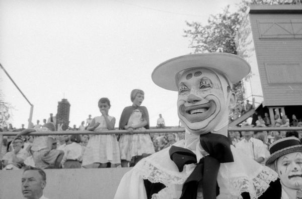 Bandleader Monty Hacker dressed in a clown suit and with smiling makeup on his face at the Shrine-Knights of Columbus benefit show at Breese Stevens Field. Another clown is standing behind him with a serious face. 