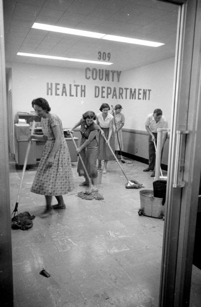 Dane County nurses and secretaries mop the floor of their third-floor public Health Department offices in the City-County Building after a faulty automatic water pump flooded the area. From left are secretaries Stella Crosse and Donna Bosenberry, nurse Barbara Kloften, and Nursing Supervisor Florence Gurholt. Also helping is Arthur Woerpel, head of the County Stores and Services Department.