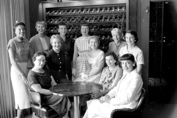 The Jaycettes are completing plans for their annual fashion show at the Memorial Union Theater. Mrs. Ben Prochaska is the general chairman of a group of eleven women. Proceeds from the show will aid the group's work for cerebral palsied and handicapped children. They are grouped in front of a floor-to-ceiling wine rack.