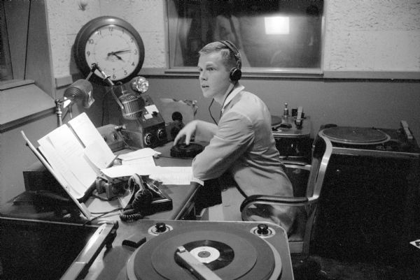 Jon Luck is shown wearing headphones, is surrounded by turntables, a microphone, and notes and scripts for commercials, while broadcasting "The Madison Hour" on WIBA.