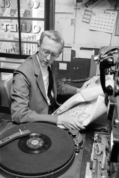 Jim Walters of WMFM holding a <i>Wisconsin State Journal</i> newspaper as he is watching a vinyl record begin. He interlaces music and comments on the news on his morning show.
