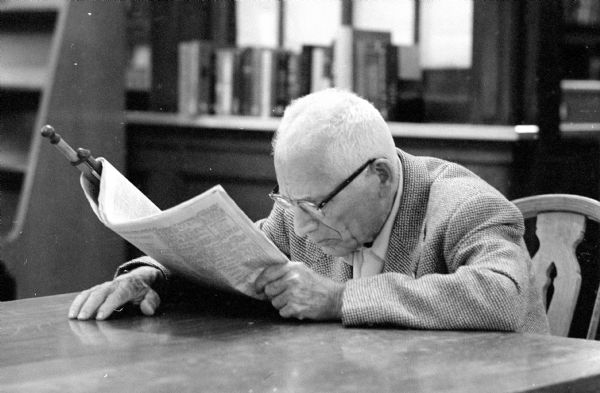 An older man wearing thick glasses and a wool blazer looks closely at a newspaper on a rod.