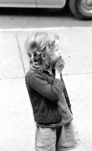 Little girl smoking a cigarette on the sidewalk.