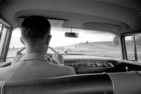 A motorist-eye view through the windshield towards a State Highway Patrol car parked beside a Wisconsin highway. The photographer, sitting in the backseat is visible in the rear-view mirror.