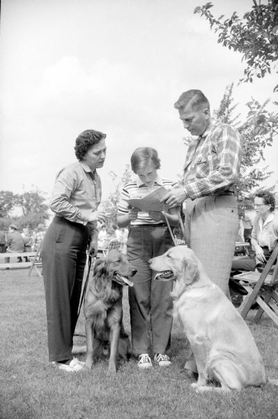 Participants in the 4th Annual Dog Obedience Trials held at the Double A Arena near Cambridge look over the event program. Shown are Mr. and Mrs. Richard Harper (4318 Odana Road) with their daughter Janet (age 10). Their two Golden Retrievers, Skipper and Buck, look on.   