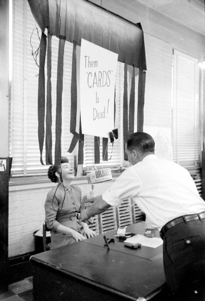 <i>Wisconsin State Journal</i> staff reporter, June Dieckmann seated at a desk. Behind her on the wall is a poster saying "Them Cards is Dead." The poster refers to the Milwaukee Braves winning the National League baseball championship over the St. Louis Cardinals. A man in a white shirt is leaning over her desk to hand her a pistol as a photo prop.
