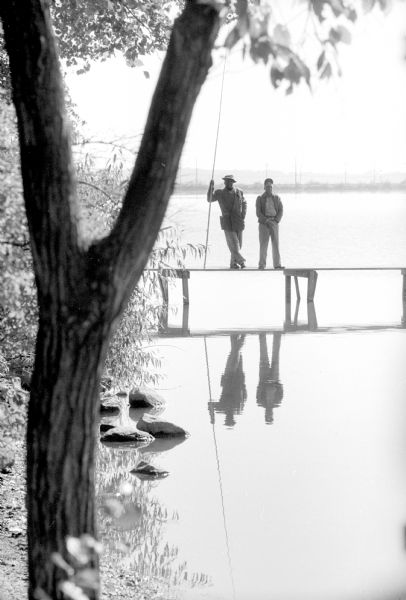 Two fishermen reflected in Monona Bay while standing on a dock. One man is wearing a hat and leaning on a long  fishing pole, and the other man is standing beside him. A shoreline is on the horizon. A tree frames the foreground edge. Sailboat masts are silhouetted in the background.
