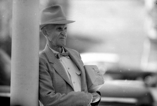 A man holding a paper-wrapped package under his arm pauses while shopping to watch the demolition of the Pioneer block and Madison Theater on the Madison Capitol Square. The buildings are to be replaced with a new building housing a J.C. Penney store. The photograph was taken with a new telephoto lens.