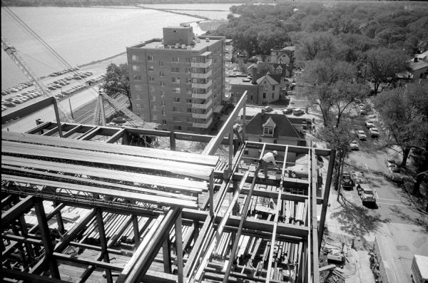 Elevated view of the West Tower of the State Office Building at 1 West Wilson Street while it is under construction. Steel vertical and horizontal members, and a construction worker are in the foreground, with Lake Monona and Monona Bay in the background. 