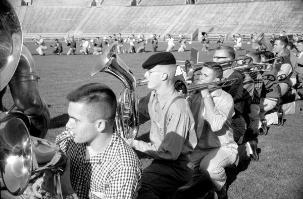 Members of the U.W. marching band practicing the half-time show prior to the U.W. vs. Marquette football game. They are in formation in the center of the grass field at Camp Randall stadium. Tuba and trombone players are in the foreground. 