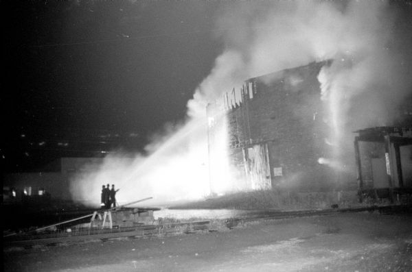 Three firemen are spraying water on a fire at the abandoned North Western Railway ice house, located on South Brearly Street along the railroad tracks between South Brearly and Ingersoll Streets. Arson was suspected, caused by a gang of teenagers roaming the area after the Madison Central High football game against Kenosha High School at Breese Stevens Field.      