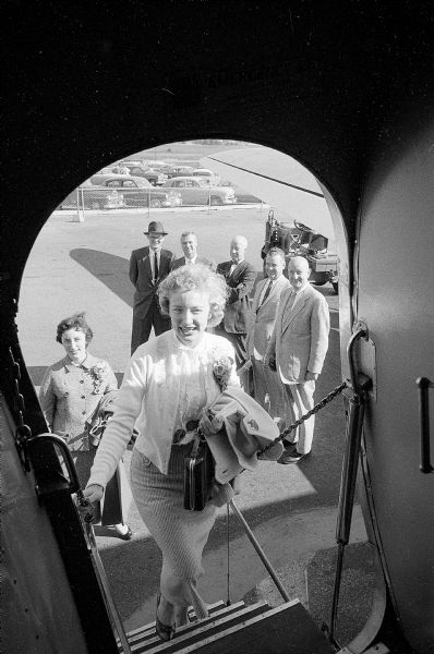 Carol Laufenberg, a Madison law firm secretary, boarding a plane headed for the University of Notre Dame where she will be queen of the Arts and Letters Ball. Behind her is Mary Kinsella, a friend who will also attend the dance. Lined up to bid her farewell are (left to right): Madison lawyers Paul Gartzke, John L. Bruemmer, Randolph Conners, Earl Cooper, and Edward J. Owens. 