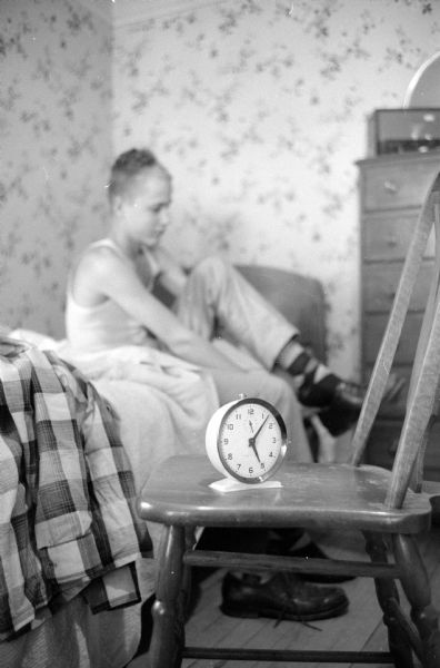Newspaper boy Malcolm Bourne, 113 Lathrop Street, dressing before dawn  to prepare for completing his daily newspaper route. The hands on his alarm clock in the foreground read seven minutes past five. He is sitting on his bed in the background by a dresser putting on his shoes. 