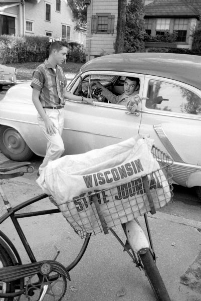 Newspaper delivery boy Mathew Bourne, 113 Lathrop Street, checks-in with his <i>Wisconsin State Journal</i> district manager Clint Vondoll, who is sitting in his car in the road. Bourne's bike is on the sidewalk in the foreground with his carrier bag.