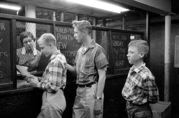 Brothers Malcolm, Mathew and Sidney Bourne, 113 Lathrop Street, line up to pay their bill for copies of the <i>Wisconsin State Journal</i> that they deliver daily. Patricia Sadler is sitting behind the cage at the newspaper office. 