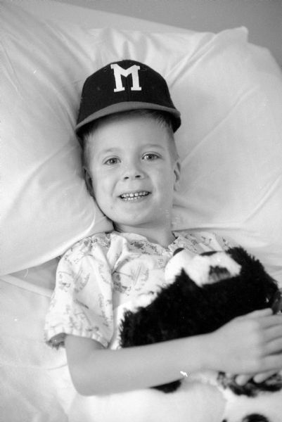 Jeffrey McVeigh, age 5 from Green Bay, recovering from a five-hour operation at University Hospital to repair a hole in the inner walls of his heart. He is laying a hospital bed smiling while holding a stuffed animal. He is wearing a baseball cap with the letter "M" on it. 