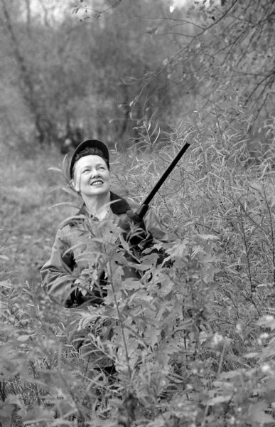 Anne Marshall Kleist, a successful owner of the women's clothing store Kleist and Wiggs, enjoying the sport of hunting. She is holding a shotgun while crouching behind some shrubs. 