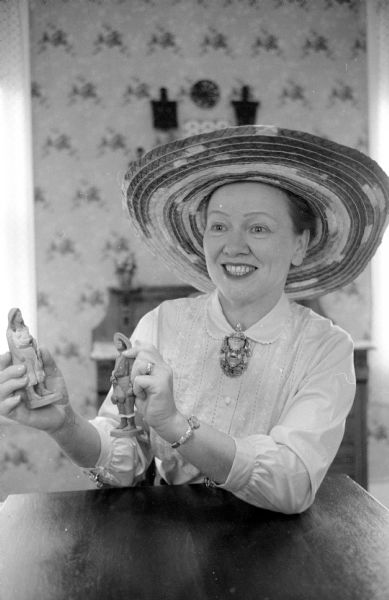 Anne Marshall Kleist in her store Kleist and Wiggs showing off merchandise from Mexico. She is wearing a sombrero on her head, and is holding two small statues. 