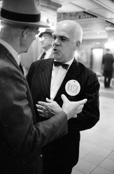 Phileo Nash (facing camera with a name button on his lapel) seeks re-election as State Democratic Party Chairman at the State Democratic Convention held at the Hotel Loraine. He is shown "buttonholing" a convention delegate in the hotel lobby.   
