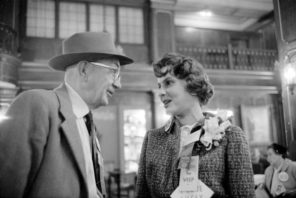 Jim Barry of Leona greets Ellen Proxmire, wife of Democratic Senator William Proxmire, at the Democratic Party State Convention inside the lobby of the Hotel Loraine. Mrs. Proxmire has a corsage on her lapel, and political badges on her coat.