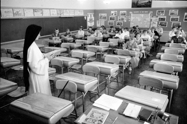 Vacant desks at St. Bernard Catholic Grade School, located at 2438 Atwood Avenue, are typical due to a flu outbreak among city pupils in Madison. Students are seen in desks towards the back of the room, and the Sister is instructing at the front.