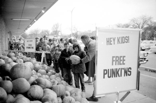 "Free punki'ns" are being given to children by the Madison Sears store and the Y Men's Club of the YMCA. The pumpkins are to be carved and entered in a contest at the City YMCA. William Simms (right), club member, is helping to hand them out.