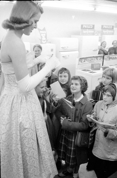 Young Madison girls requesting autographs of Miss America, Marilyn Elaine van Derbur, during a fashion show at the Casey and O'Brien, Inc. store on Atwood Avenue. She was in Madison on a stop of her tour of the country. She is wearing a tiara, long white gloves and a gown. Philco appliances are in the background.