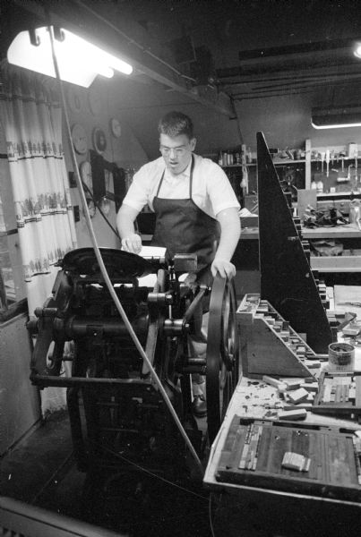 Young John Schindler, 18, the son of Dr. Schindler, standing and wearing an apron behind a printing press that he uses to do "job" printing work for his business, as he has done since age 12.