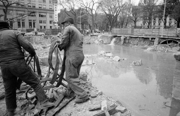 Two workmen in coveralls are laying hose to pump water from a flooded pit where the Pioneer Building was demolished at Main Street and Monona Avenue. On the right is the broken 4-inch pipe gushing water in the site. In the upper left corner is the Bank of Madison at 1 West Main Street.