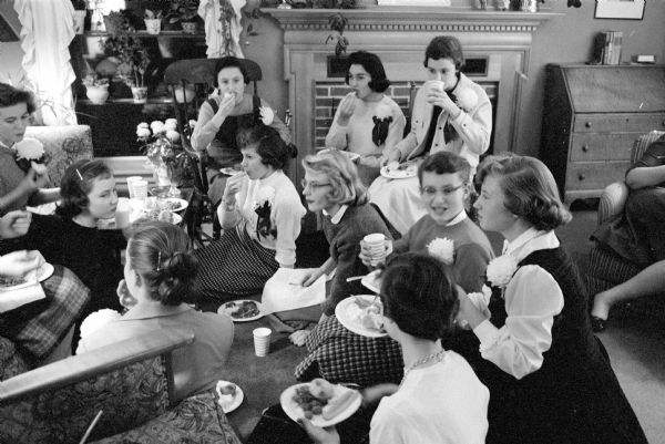 Edgewood High School teen-age girls gather for a potluck before a football game. The potluck was at the home of Mark and Mary Schmitz  and hosted by their daughter, Martha. The girls are crowding in the living room. Some are wearing chrysanthemum corsages on their sweaters.