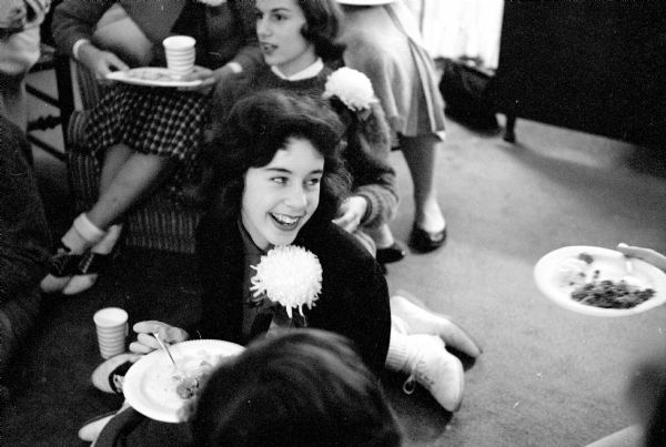 Carol Tobin enjoying the teen-age girls' potluck before the Edgewood High School football game. The potluck was at the home of Mark and Mary Schmitz and hosted by their daughter, Martha. The girls are gathered in the living room, sitting on the floor and chairs. Some are wearing chrysanthemums for the occasion.