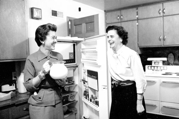 Two of the mothers show amusement in the kitchen while helping out at the teen-age girls' potluck before an Edgewood High School football game. Mary Schmitz (left, mother of the daughter who is hosting) shared a moment with Virginia Rogers as she takes a pitcher of milk from her refrigerator.