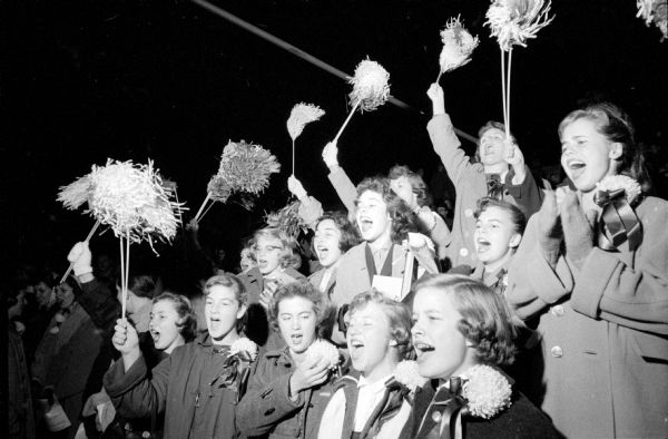 Girls cheer on the Edgewood High School football team after having their pre-game potluck at the home of Mark and Mary Schmitz. They are seen cheering and raising pom-poms. Some are wearing chrysanthemum corsages for the occasion.