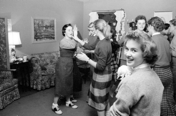 A group of teen-agers enjoying themselves at the pre-game potluck before the Edgewood High School football game. The potluck was at the Mark and Mary Schmitz home and was hosted by their daughter, Martha. A couple girls are playing patty cake in the living room. Some are wearing chrysanthemum corsages for the occasion.