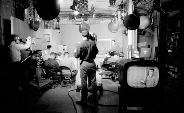 Television broadcast of a University of Wisconsin class by Professor M. Robert Irwin. Cameramen are standing at recording equipment and at installed lighting beams down on the studio classroom. The professor is seen at the chalkboard on a tv monitor in the right foreground.